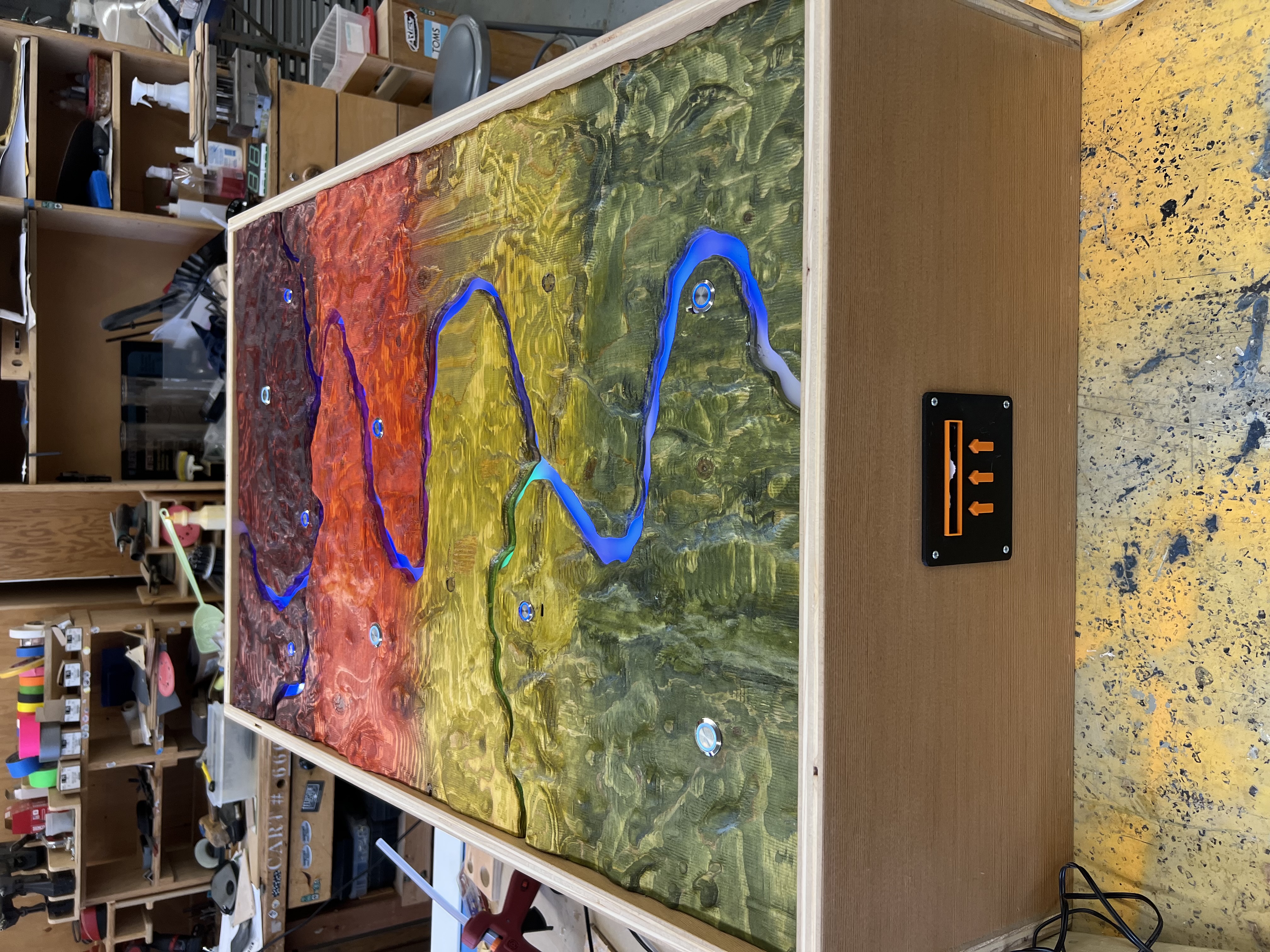 a wooden box, the top is color coded by height with buttons and streams of leds, a slot for a reciept paper to be printed is visible on the front side of the box.